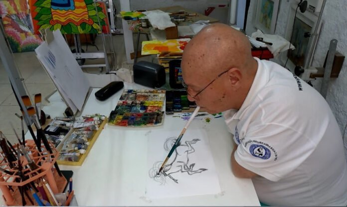 Descrição da imagem #pracegover: Foto do artista Marcos Paulo durante a pintura de um quadro. Ele não tem os braços e está pintando uma tela com a boca. Fim da descrição.
