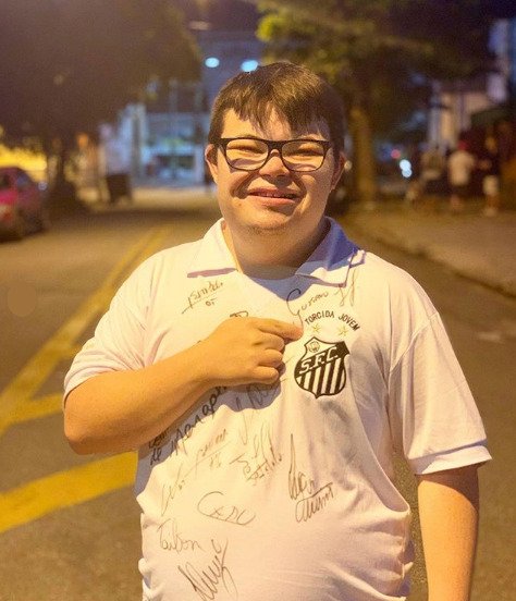 Descrição da imagem #pracegover: Foto do Casis Neto. Ele está na rua e veste uma camisa autografada do Santos. Casis é um jovem com síndrome de Down. Ele é moreno, tem cabelos castanhos, lisos, e usa óculos. Fim da descrição.