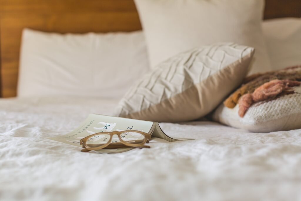 Descrição da imagem #pracegover: Uma cama confortável. Sobre ela estão travesseiros e almofadas, além de um óculos e um livro. Fim da descrição.