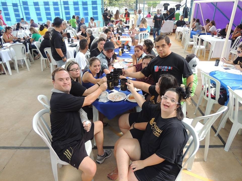 Descrição da imagem #pracegover: Foto do grupo de jovens com síndrome de Down durante encontro do Felicidade Down. Eles estão sentados em uma mesa e brindam. Todos usam uma camiseta preta. Fim da descrição.