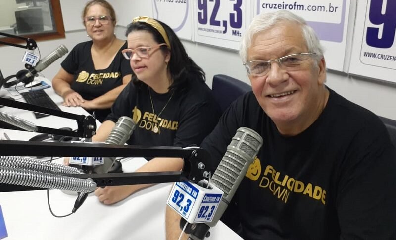 Descrição da imagem: foto de Tomás e Celia, fundadores do Felicidade Down, com a filha Bruna. Eles vestem uma camiseta preta e estão em uma rádio. Fim da descrição.