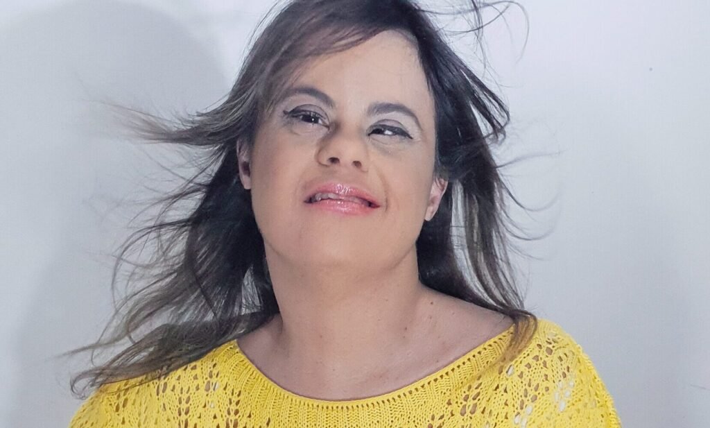 Descrição da imagem #pracegover: Foto da Fernanda Honorato. Ela é uma mulher jovem, morena, tem cabelos lisos longos e castanhos. Ela está usando uma blusa rendada amarela e calça preta. Fernanda faz pose para a foto. Ela tem síndrome de Down. Fim da descrição.