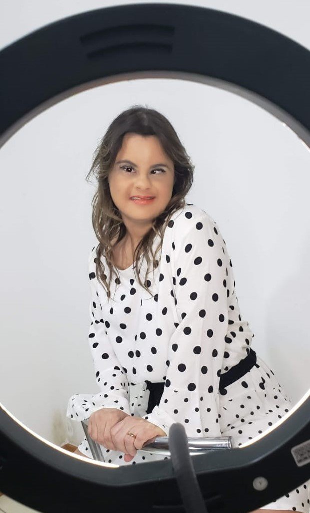 Descrição da imagem #pracegover: Foto da Fernanda Honorato. Ela é uma mulher morena, tem cabelos lisos longos e castanhos. Ela está usando uma blusa branca com bolinhas pretas e faz pose para a foto. Fernanda tem síndrome de Down. Fim da descrição.