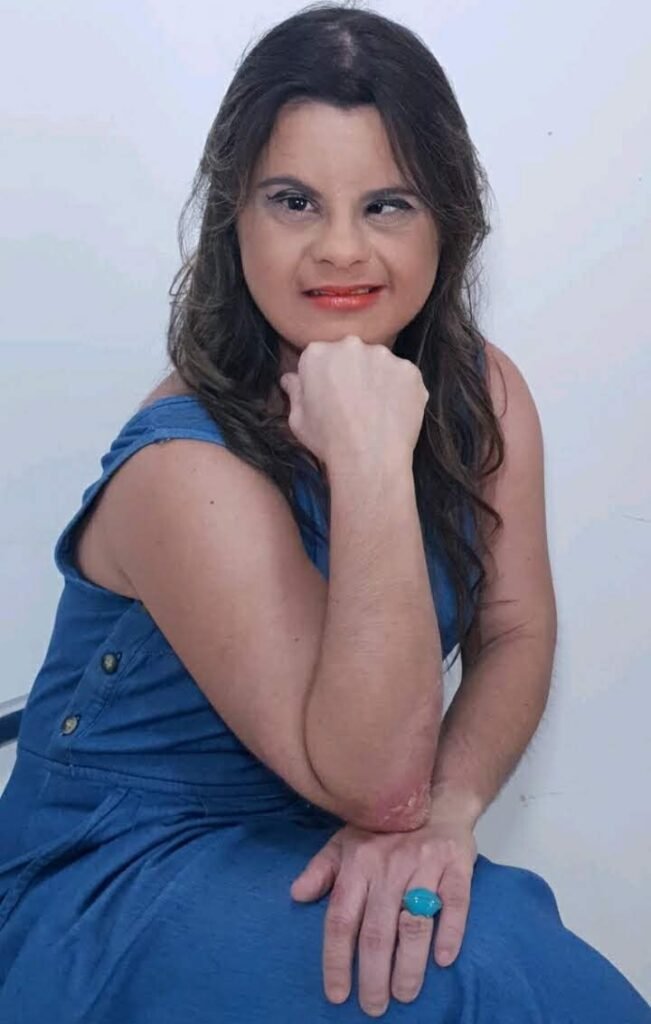 Descrição da imagem #pracegover: Foto da Fernanda Honorato. Ela é uma mulher morena, tem cabelos lisos longos e castanhos. Ela está usando um vestido jeans e faz pose para a foto. Fernanda tem síndrome de Down. Fim da descrição.