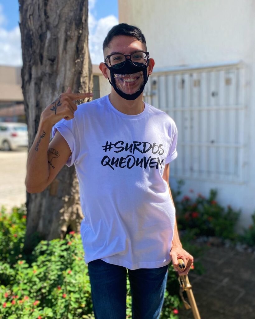 Descrição da imagem #pracegover: Ivan Baron está usando uma camiseta branca e calça jeans. Nela está escrito: Surdos que ouvem. Ivan está apontando para sua máscara na cor preta com a parte frontal transparente. Fim da descrição.