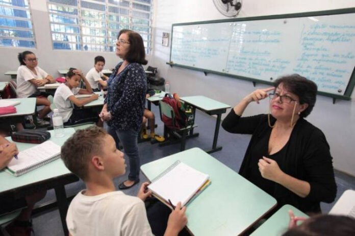 Descrição da imagem #pracegover: Foto de uma sala de aula. A professora está no centro da sala e, em primeiro plano, vemos um estudante sentado em sua carteira. À sua frente está uma intérprete de Libras. Fim da descrição.