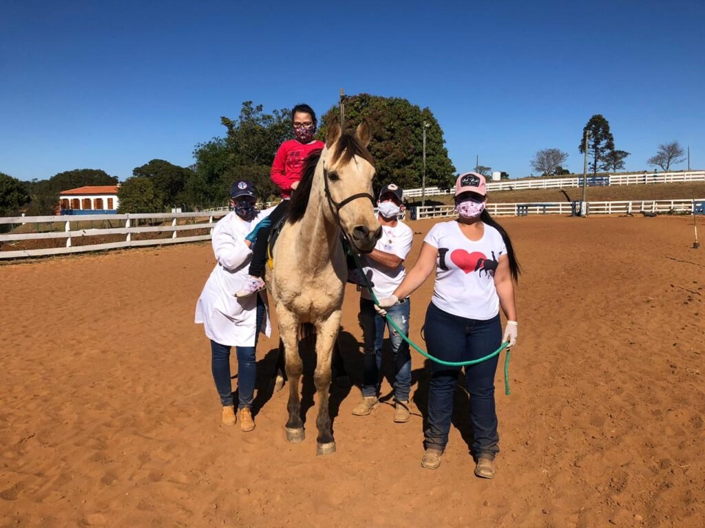 Descrição da imagem #pracegover: Uma menina está montada no cavalo e três profissionais de equoterapia estão no chão, ao seu lado. Fim da descrição.