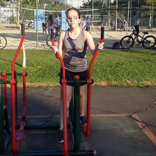Descrição da imagem #pracegover: Sara está praticando exercícios em um parque. Ela é uma moça loira e está com os cabelos presos. Sara veste uma regata cinza e um shorts preto. Fim da descrição.