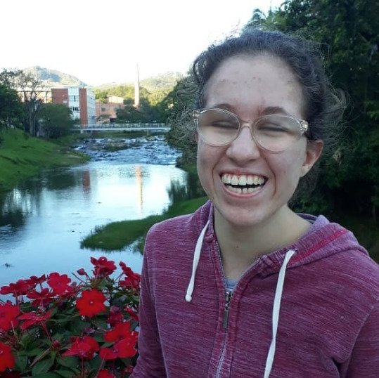 Descrição da imagem #pracegover: Sara está em um parque. Ela é uma moça loira e está com os cabelos presos. Sara veste um casaco de capuz cor de rosa. Atrás dela está um rio com pequenas flores vermelhas em destaque. Atrás dela também estão plantas e pedras em meio às águas do rio. Sara está sorrindo. Fim da descrição.