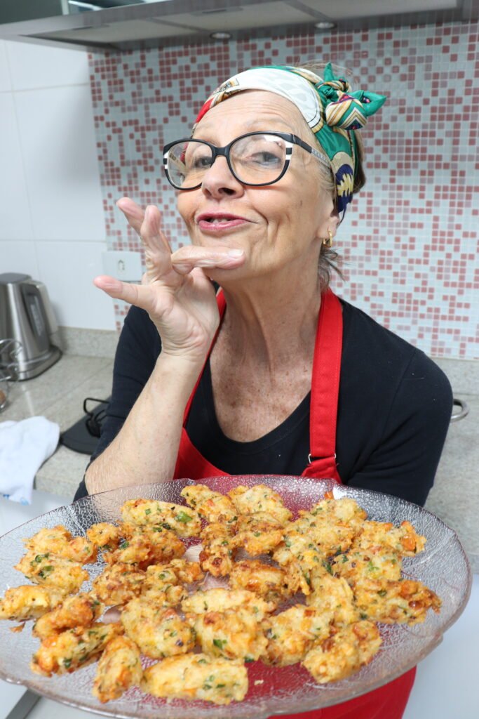 Descrição da imagem #pracegover: Foto da Vera, idealizadora e apresentadora do canal Cozinhando em Libras. Vera está na cozinha. Ela segura uma bandeja com bolinhos de arroz. Vera é uma mulher morena. Ela usa óculos e está vestindo uma blusa preta e um avental vermelho. Ela está com uma bandana estampada com predominância da cor verde, nos cabelos. Vera faz um sinal em Libras. Fim da descrição.