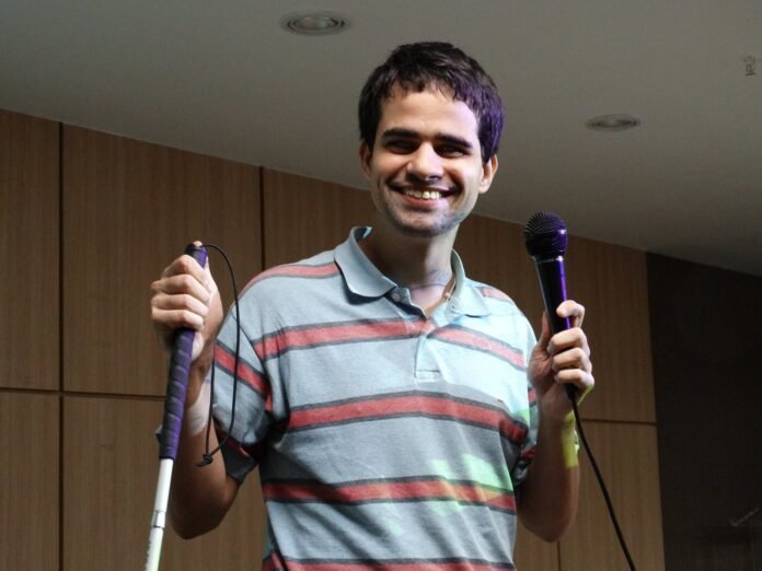 Descrição da imagem #pracegover: Foto de Marcos Lima. Ele é um rapaz moreno e tem cabelos castanhos. Marcos veste uma camisa polo azul com listras e segura nas duas mãos bengalas. Marcos está sorrindo. Fim da descrição.