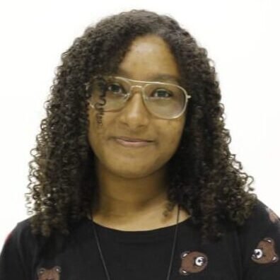 Descrição da imagem #pracegover: Retrato da estudante de jornalismo, Andressa Franco. Ela é uma moça morena e tem cabelos cacheados, compridos. Andressa usa óculos de grau e sorri. Ela veste uma camiseta preta com estampas de ursinhos. Fim da descricão.