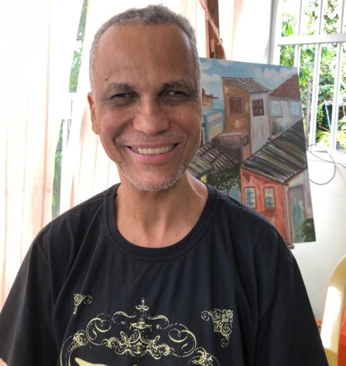 Descrição da imagem #pracegover: Retrato de Edinilson Sacramento. Ele é um homem moreno de cabelos grisalhos e curtos. Ednilson usa uma camiseta preta e está sorrindo. Ao fundo vemos uma janela e um quadro, que está em um cavalete. Fim da descricão.