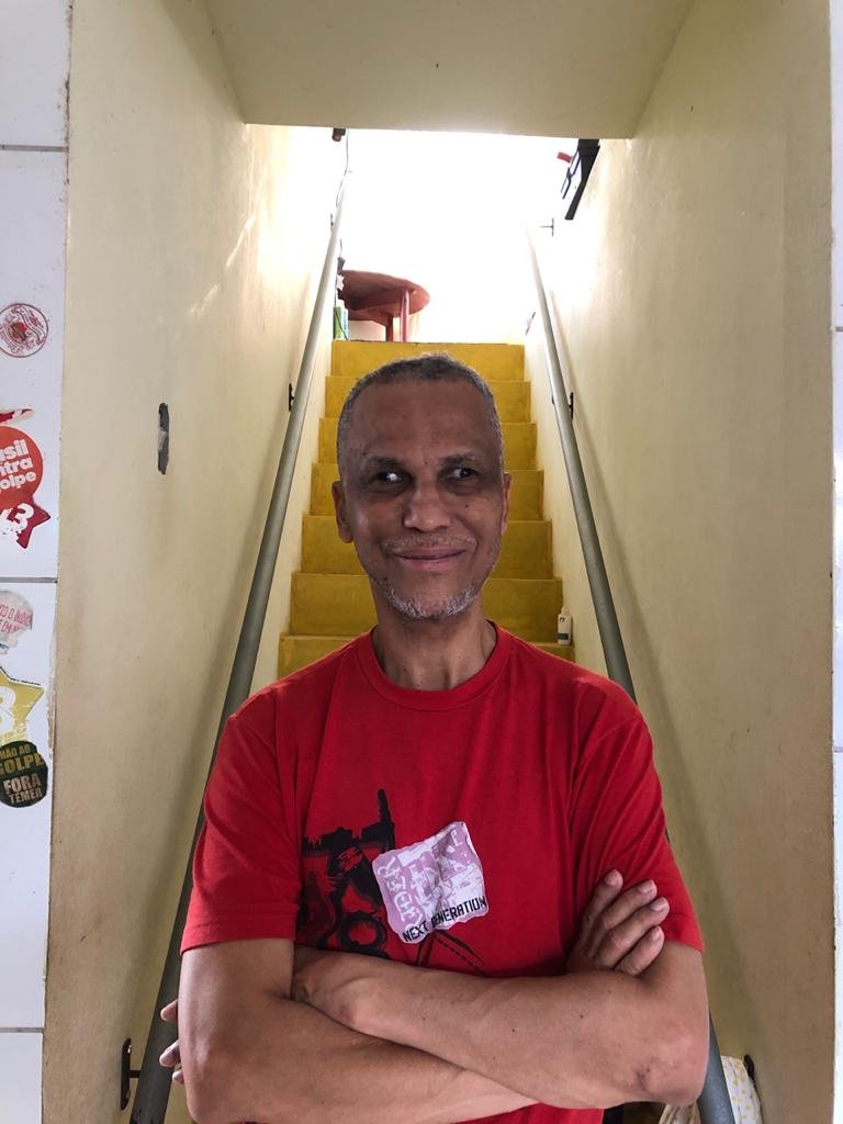 Descrição da imagem #pracegover: Retrato de Edinilson Sacramento. Ele é um homem moreno de cabelos grisalhos. Ednilson usa uma camiseta vermelha, está com os braços cruzados, e sorri. Ao fundo vemos uma escada. Fim da descricão.