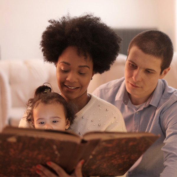 Descrição da imagem #pracegover: Foto de um casal lendo para uma criança. Fim da descrição.