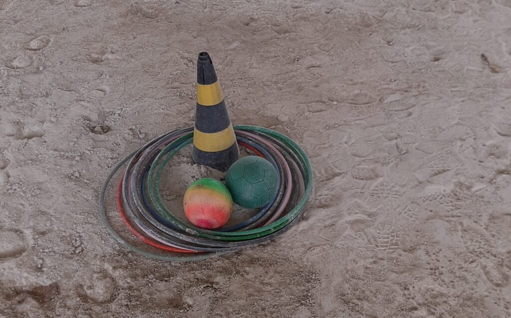 Descrição da imagem #pracegover: Foto de um cone com vários bambolês e uma bola, no setting terapêutico, que tem chão de areia. Fim da descrição.