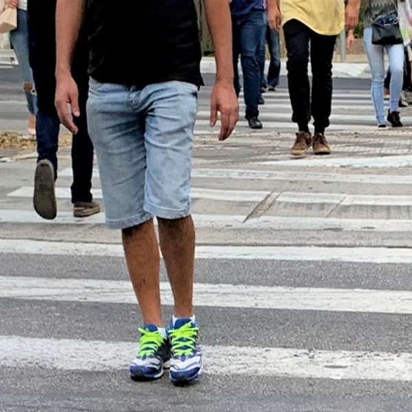 Descrição da imagem #pracegover: Foto de pessoas atravessando uma faixa de pedestre. Em primeiro plano, está um rapaz que veste camiseta preta, bermuda jeans e tênis com cadarço verde. Fim da descrição.