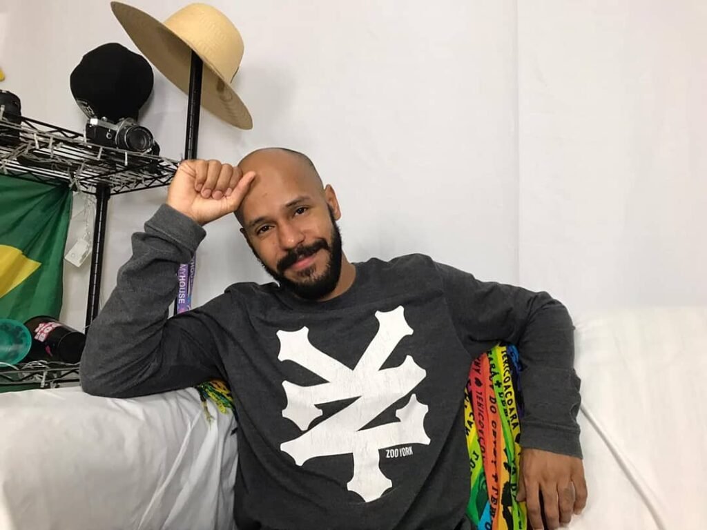 Descrição da imagem #pracegover: Foto do dançarino Maycon Calasancio. Ele está sentado em um sofá e tem a mão direita na cabeça. Maycon é um homem negro. Ele é careca e usa barba. Ele está vestindo uma camiseta preta com faixas brancas. Fim da descrição.