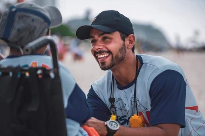 Descrição da imagem #pracegover: Foto do Patrick Souza durante evento esportivo. Ele está abaixado em frente a uma pessoa na cadeira de rodas. Ele está vestindo uma camiseta branca com mangas azuis e um boné azul. Patrick é um homem moreno e tem barba. Fim da descrição.