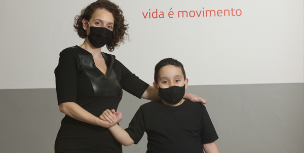Descrição da imagem #pracegover: Foto da Mariana com seu filho Dudu. Eles são os personagens símbolos do Teleton 2020. Mariana tem amputação bilateral e Dudu tem mielomeningocele. Ambos realizam tratamento na AACD. Eles estão lado a lado e usam roupas preatas. Fim da descrição.