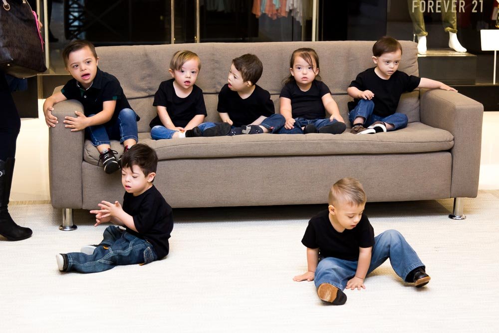 Descrição da imagem #pracegover: Foto de sete crianças com síndrome de Down. Algumas estão sentadas em um sofá e outras estão sentadas no chão. Todas elas usam camiseta preta e calça jeans. Fim da descrição.