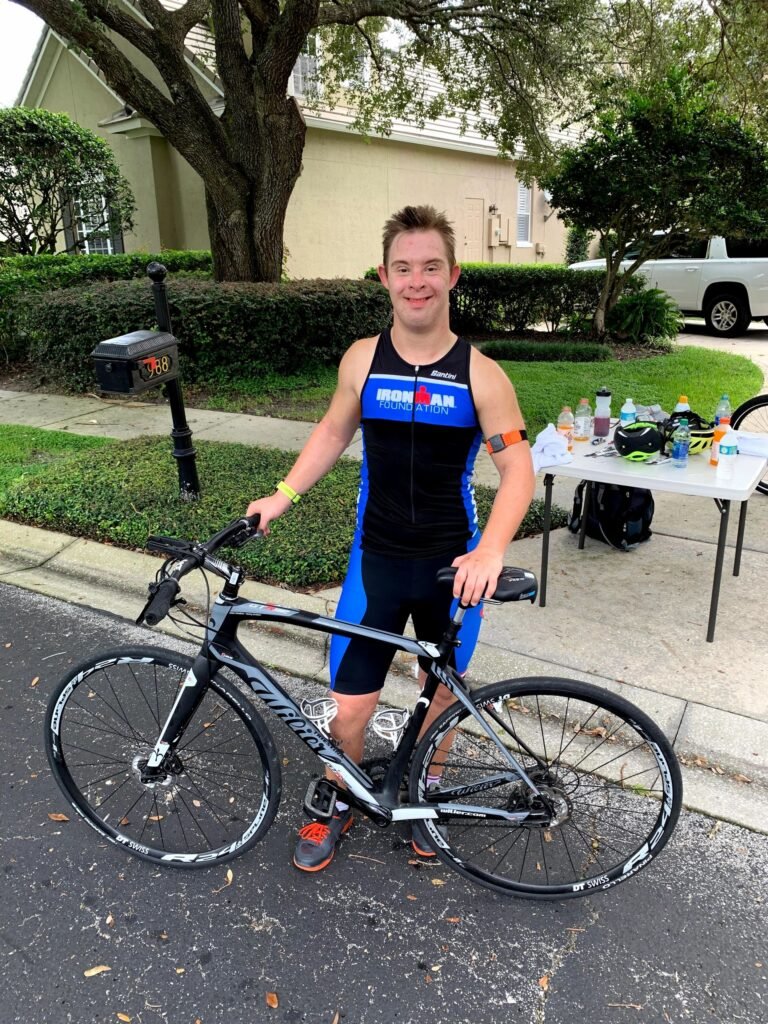 Descrição da imagem #pracegover: Foto de Chris Nikic com sua bicicleta. Chris é um jovem moreno e tem síndrome de Down. Ele usa uma roupa de ginástica preta e azul. Fim da descrição.