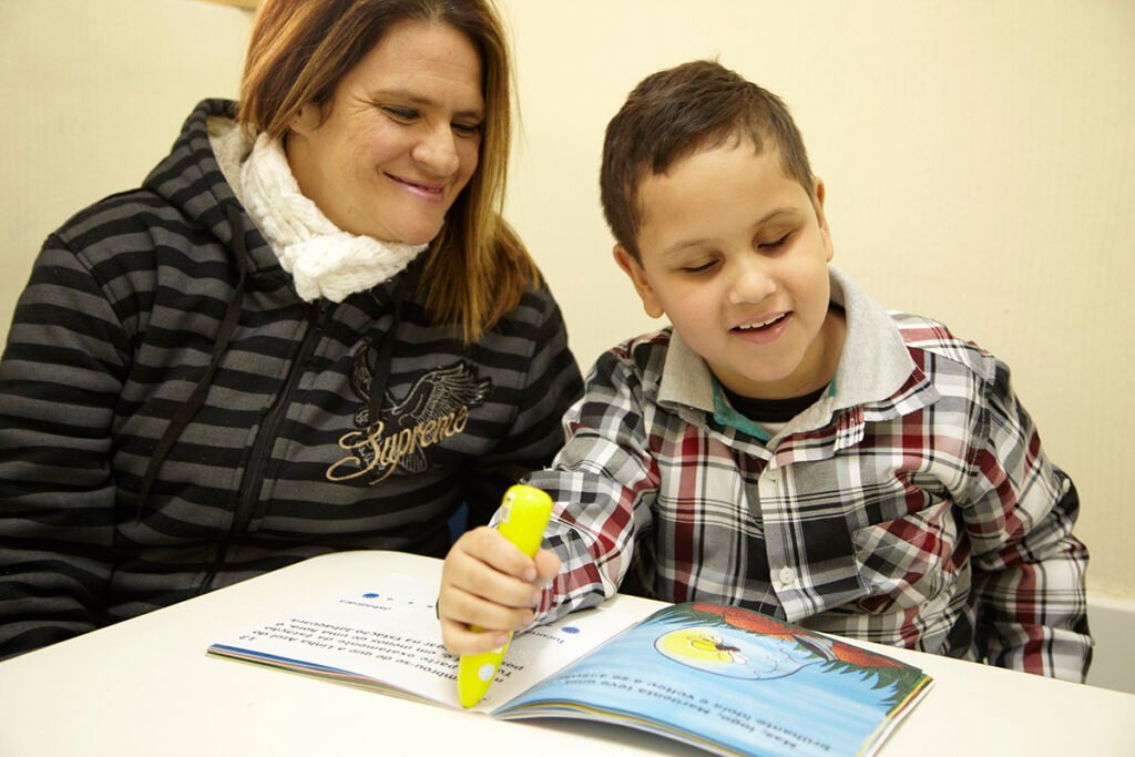 Descrição da imagem #pracegover: Foto de um menino cego realizando uma atividade em um livro adaptado. Ele é moreno e tem cabelos castanhos curtos. O menino está sorrindo e ao seu lado está uma mulher. Fim da descrição.