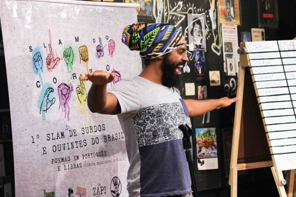 Descrição da imagem #pracegover: Foto de uma apresentação do Slam do Corpo. Um homem está com os braços abertos. Ele tem um turbante na cabeça. Fim da descrição.