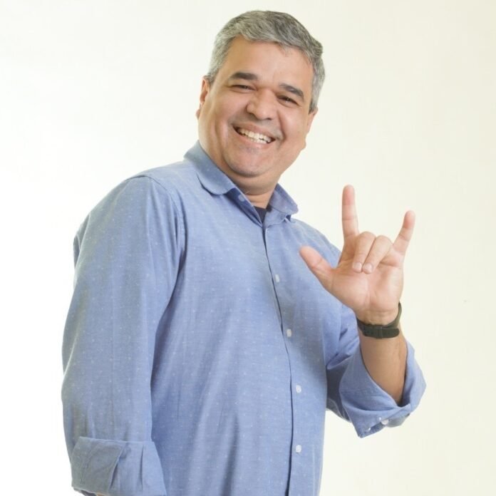 Descrição da imagem #pracegover: Foto de Marcelo Bessa. Ele é um homem moreno, de cabelos curtos e grisalhos. Marcelo está vestindo uma camisa social azul e faz o sinal universal de I Love You, em Libras. Fim da descrição.