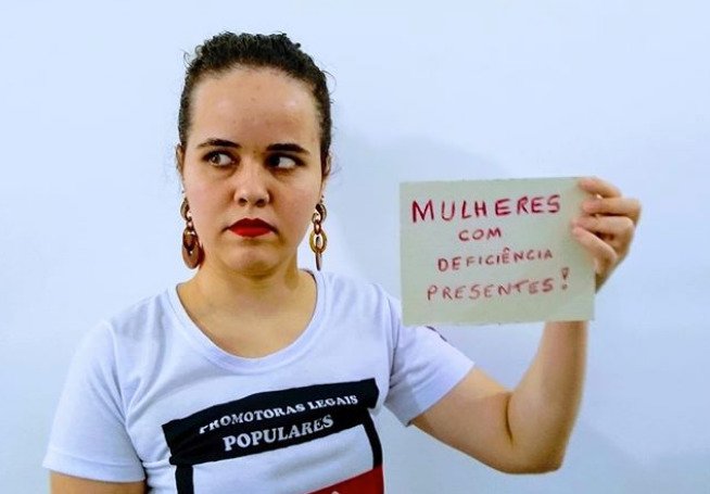 Descrição da imagem #pracegover: Mayra está com os cabelos presos em coque. Ela usa batom vermelho e veste camiseta branca com um desenho em preto branco e vermelho escrito Promotoras Legais Populares Ribeirão Preto-SP. Mayra segura um cartaz com os dizeres: MULHERES COM DEFICIÊNCIA PRESENTES. Fim da descrição.