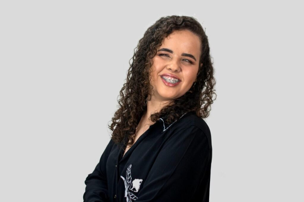 Descrição da imagem #pracegover: Mayra está de lado e tem os braços cruzados. Ela é uma mulher morena de cabelos cacheados e compridos. Mayra está usando um terninho preto e sorri. Fim da descrição.