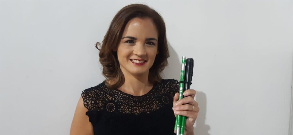 Descrição da imagem #pracegover: Foto de Priscilla Selares. Ela é uma mulher morena de cabelos castanhos na altura dos ombros. Priscilla está usando uma blusa preta e sorri. Ela está segurando sua bengala verde. Fim da descrição.