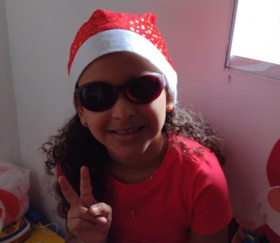 Descrição da imagem #pracegover: Foto de uma menina. Ela é morena e tem cabelos compridos e cacheados. Ela está vestindo uma camiseta vermelha, um gorro de Papai Noel e usa óculos escuros. Fim da descrição.