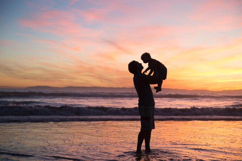 Descrição da imagem #pracegover: Foto de uma pessoa segurando uma criança no colo. Elas estão na beira da praia e ao fundo vemos o pôr do sol, em um céu alaranjado. Fim da descrição.