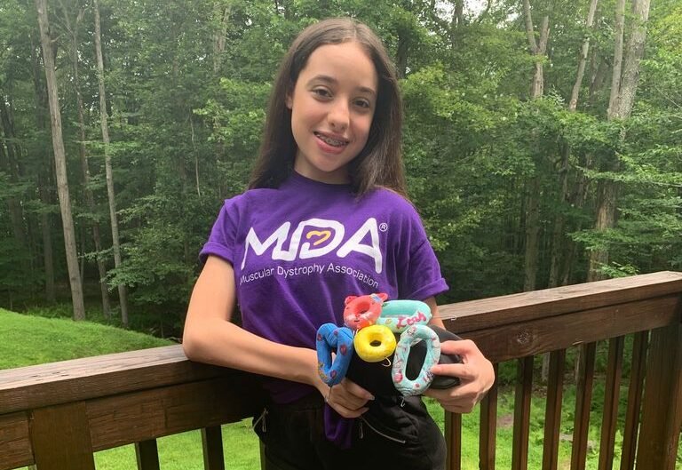 Descrição da imagem #pracegover: Foto de Leah Zelaya. Ela é uma menina morena com cabelos castanhos. Leah está vestindo uma camiseta roxa e uma bermuda preta. Ela tem próteses nas pernas e está em pé, encostada em um guarda-corpo de madeira. Leah segura algumas peças do acessório Easy Zip. Ao fundo vemos um gramado e uma área verde. Fim da descrição.