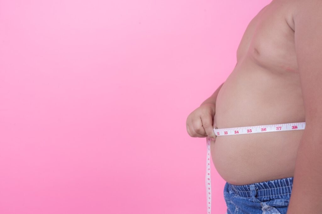 Descrição da imagem #pracegover: Foto de uma criança obesa com uma fita métrica na barriga. A imagem tem fundo cor de rosa. Fim da descrição.