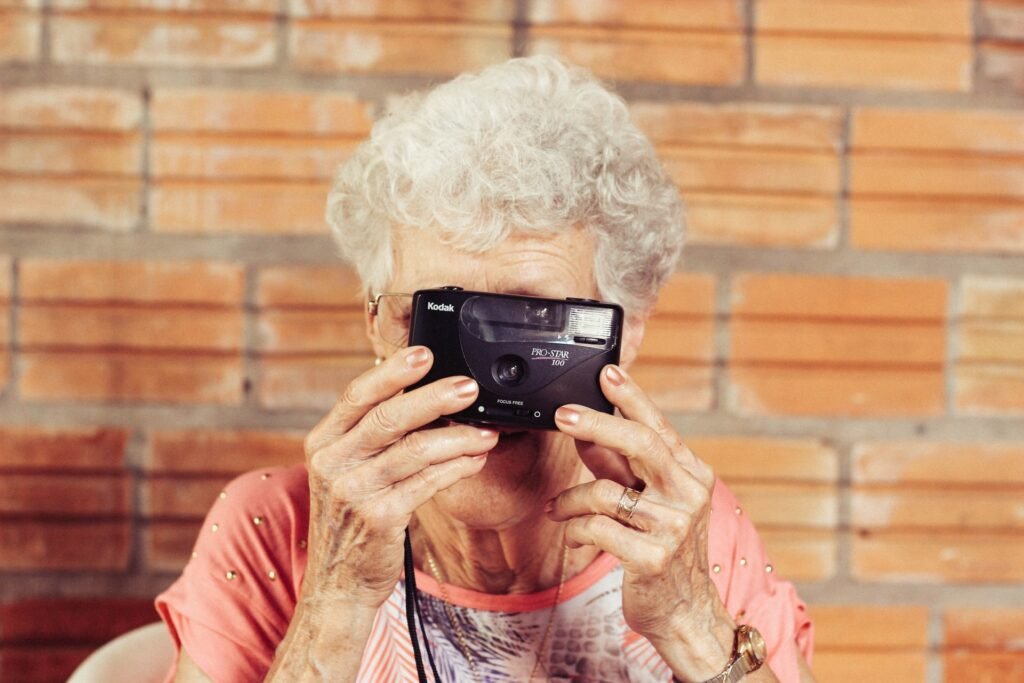 Descrição da imagem #pracegover: Foto de uma mulher idosa tirando foto. Ela está segurando uma câmera fotográfica na frente do rosto. A mulher tem cabelos brancos curtos e cacheados. Fim da descrição.