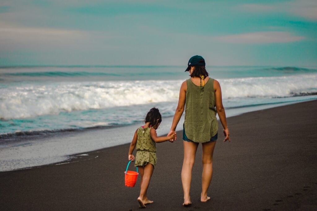 Descrição da imagem #pracegover: Foto de uma mulher e uma menina caminhando na beira da praia. Elas estão de mãos dadas e a menina segura um baldinho de areia. Fim da descrição.