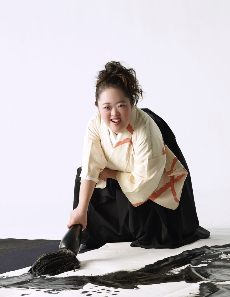 Descrição da imagem #pracegover: Shoko durante criação de uma arte. Ela segura um grande pincel e escreve sobre um tecido no chão. Shoko é uma jovem japonesa. Ela tem cabelos pretos e lisos que estão presos. Ela usa um traje japonês. Fim da descrição.
