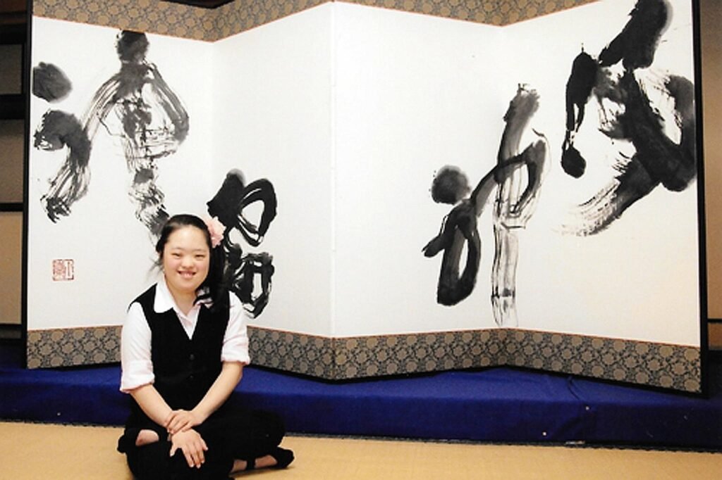 Descrição da imagem #pracegover: Shoko é uma jovem japonesa. Ela está sentada à frente de uma pintura com caligrafia japonesa. Shoko tem cabelos pretos e lisos que estão presos. Ela usa uma camisa branca, colete e calça preta. Fim da descrição.