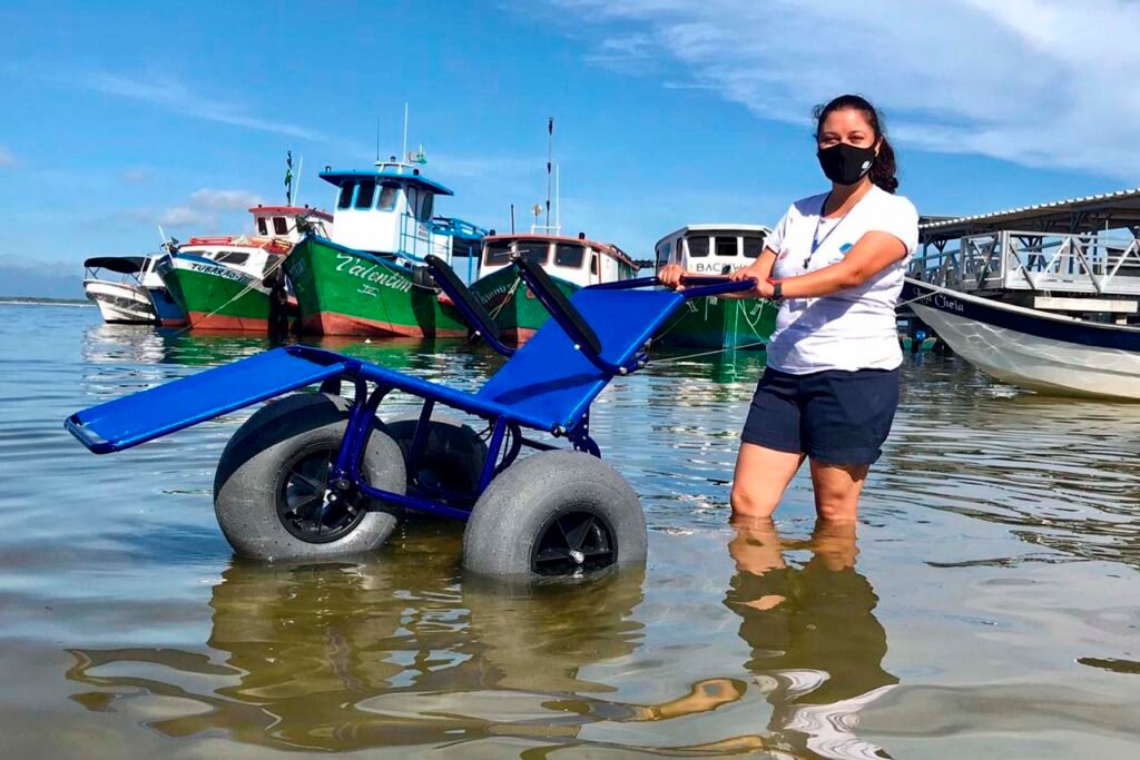 Descrição da imagem #pracegover: Foto da cadeira anfíbia. Ela tem peneus largos e estrutura azul. Uma mulher está segurando a cadeira dentro do mar. Fim da descrição.