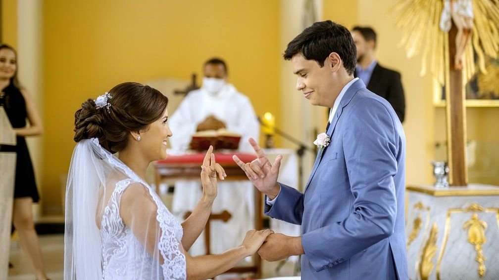 Descrição da imagem #pracegover: Os noivos estão no altar, durante a cerimônia de casamento. Eles estão fazendo o sinal de Eu Te Amo em Libras. Fim da descrição.