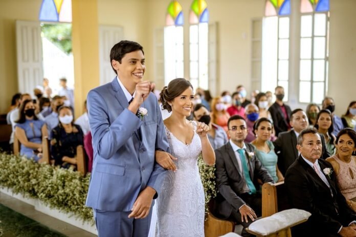 Descrição da imagem #pracegover: Os noivos no altar, durante a cerimônia de casamento. Eles estão fazendo o sinal de Sim, em Libras. Ao fundo vemos os convidados na igreja. Fim da descrição.