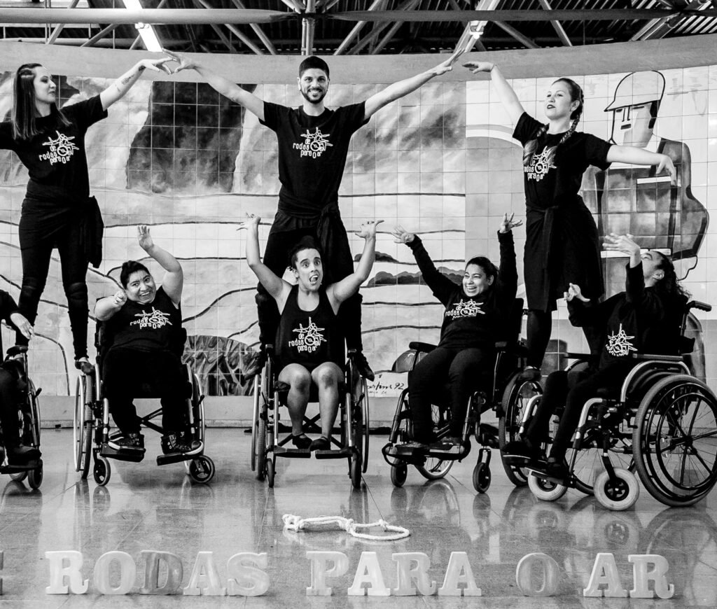 Descrição da imagem #pracegover: Foto preto e branco de uma apresentação da Cia de dança de Rodas Para o Ar. Dançarinos na cadeira de rodas em uma performance de dança. Fim da descrição.