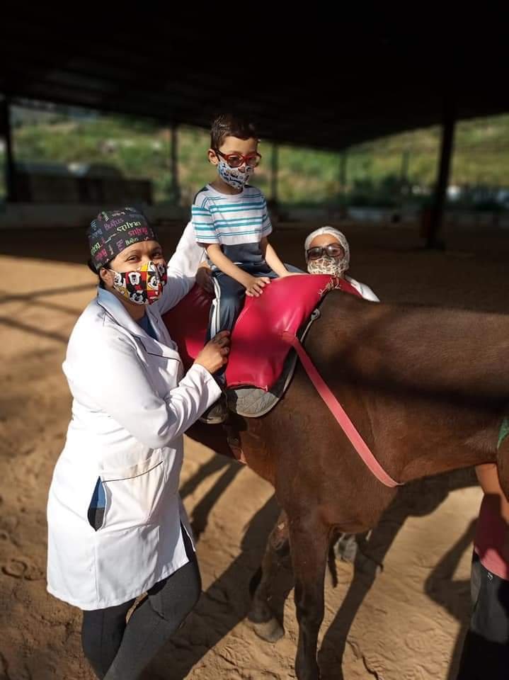 Descrição da imagem #pracegover: Uma criança está montada em um cavalo marrom. A equipe de equoterapia está ao lado do cavalo. Fim da descrição.