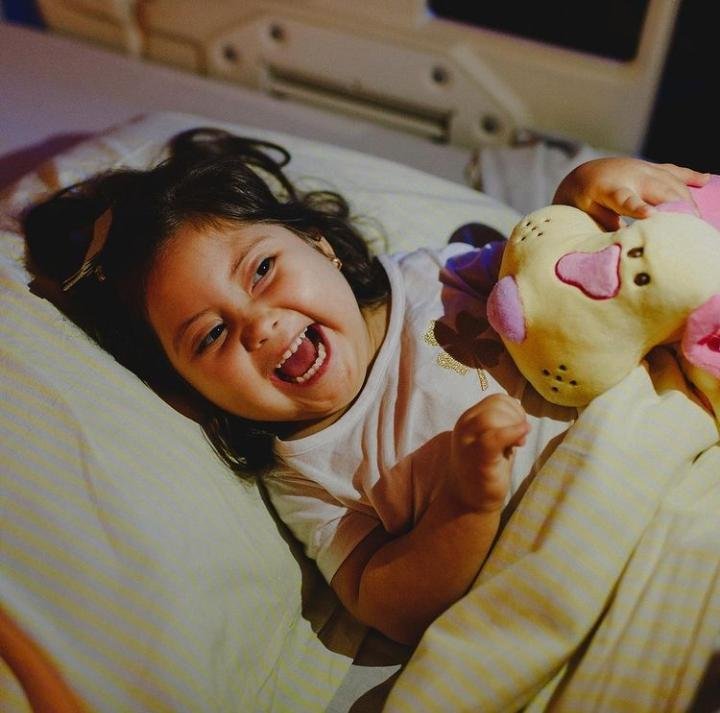 Descrição da imagem #pracegover: Foto da pequena Mel. Ela é uma menina morena e tem cabelos castanhos, lisos. Mel tem síndrome de Down. Ela está deitada na cama e sorri. Fim da descrição.