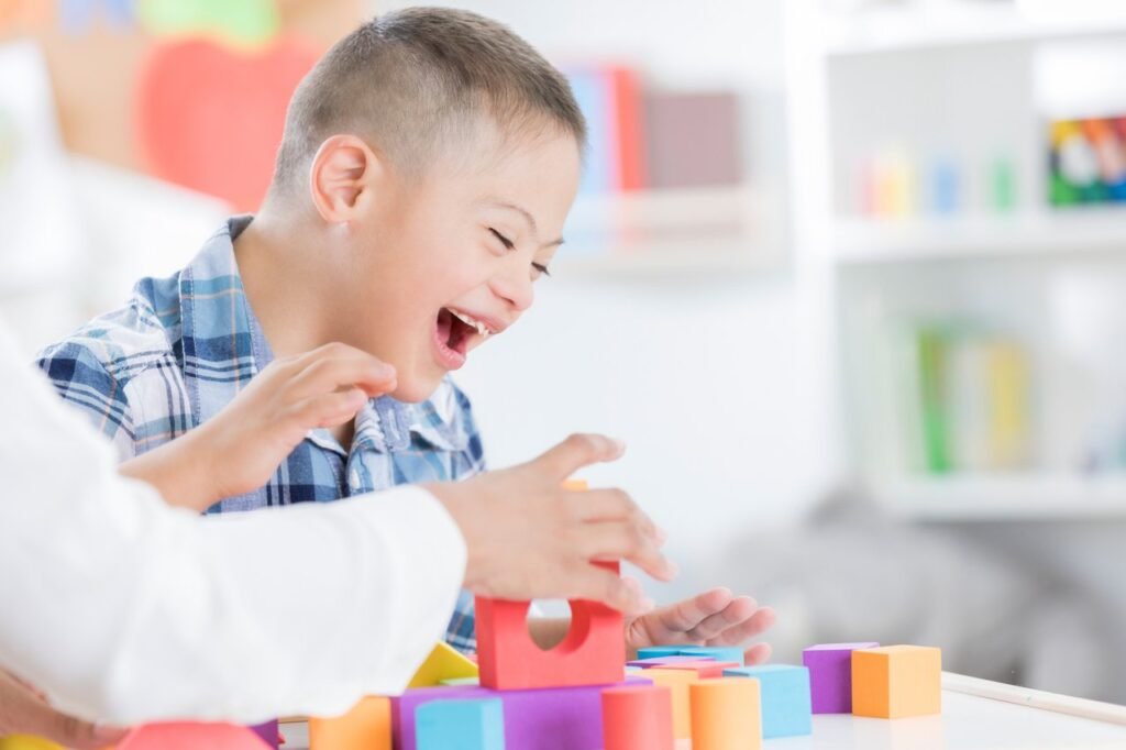 Descrição da imagem #pracegover: Foto de um menino brincando com peças coloridas sobre a mesa. O menino tem síndrome de Down. Ele está acompanhado de uma pessoa, e sorri. Fim da descrição.
