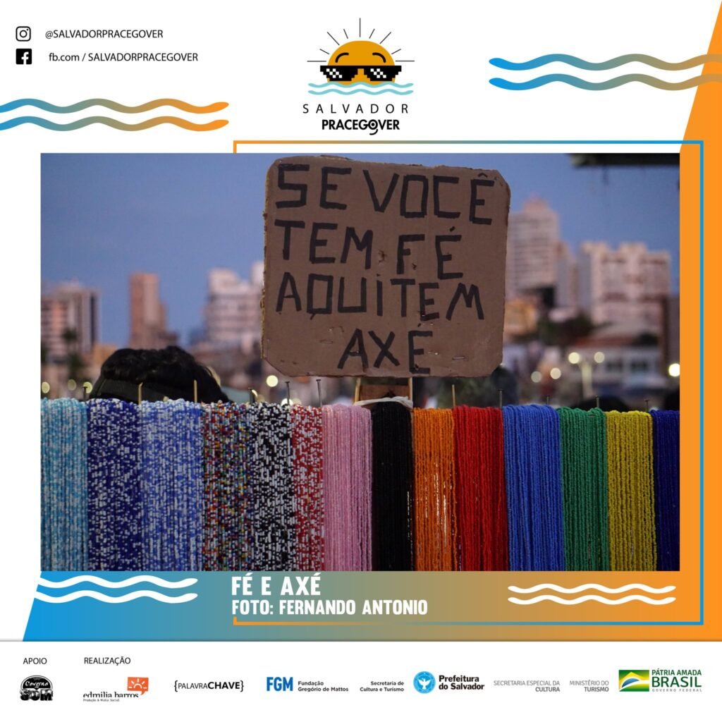 Descrição da imagem #pracegover: Foto de muitos colares de miçangas coloridas estão sobre um pedaço de madeira na horizontal. Eles estão milimetricamente organizados um ao lado do outro. Em cima, em um papelão preso numa madeira vertical, está escrito: Se você tem fé aqui tem axé. Ao fundo, prédios desfocados. Fim da descrição.