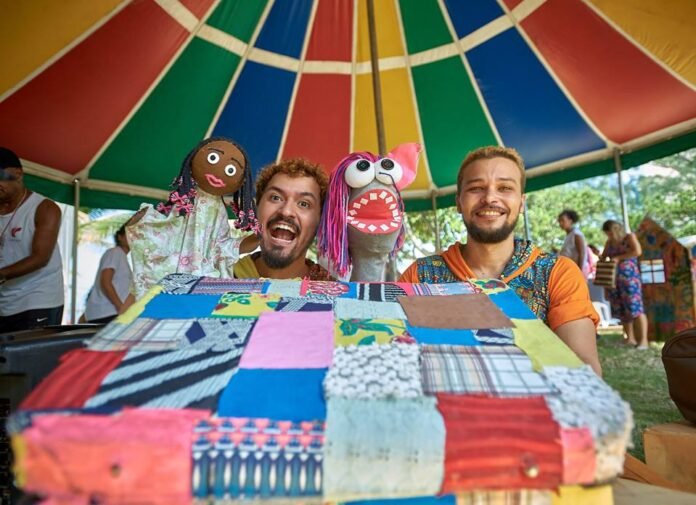 Descrição da imagem #pracegover: Foto de dois contadores de histórias com marionetes. São dois rapazes que estão em um local com tenda colorida. Eles estão com marionetes nas mãos. Fim da descrição.