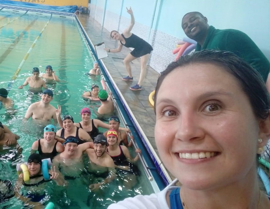Descrição da imagem #pracegover: Foto de Raphaela com atletas da natação. Ela está em primeiro plano, fora da piscina, e em segundo plano vemos os atletas dentro da piscina. Na lateral da piscina estão outros dois integrantes da equipe técnica, um homem e uma mulher. Fim da descrição.